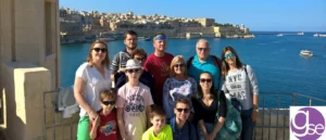 A group of teenagers and adults posing together on a waterfront viewpoint overlooking a Mediterranean harbour with historic limestone buildings
