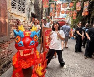 Visitors posing with a red decorative lion statue in a narrow street lined with red lanterns and traditional buildings